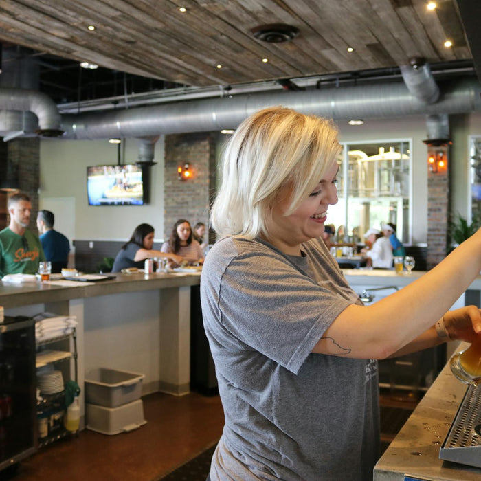 A smiling bartender pours a beer for customers in a lively, industrial-style bar setting.