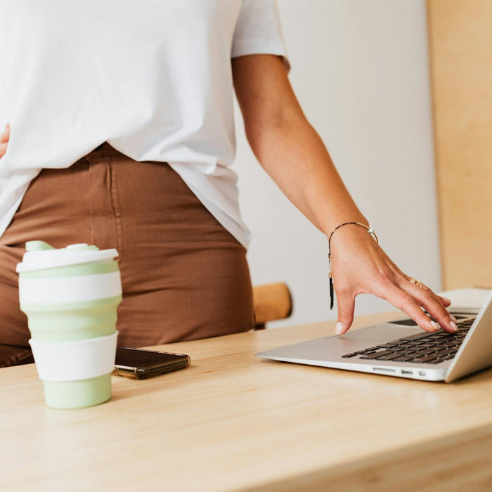 A woman stands by a wooden desk using a laptop with a reusable cup nearby. Modern lifestyle concept.