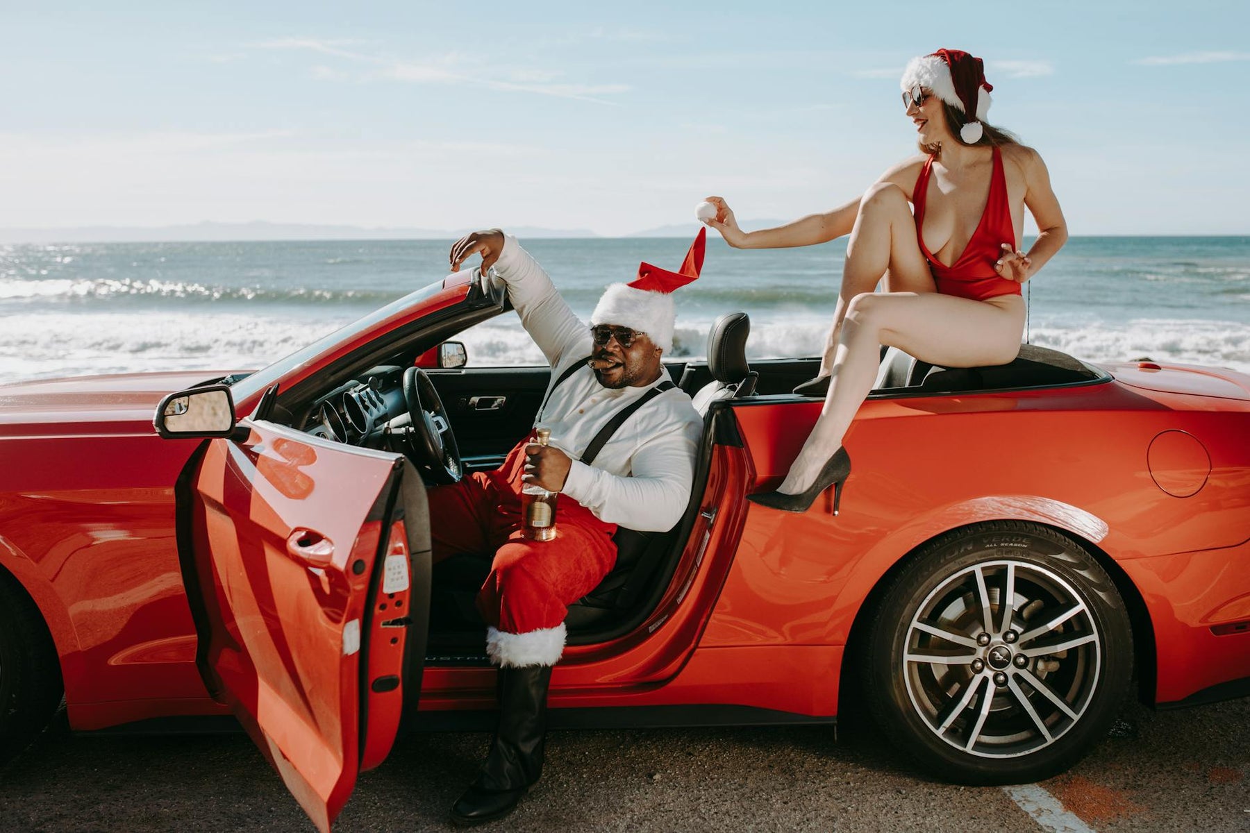 Santa Claus and woman in Santa hats enjoying a convertible by the seaside, showcasing a festive holiday scene.