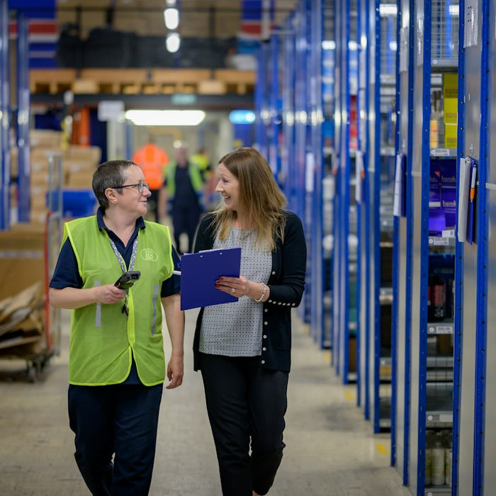 Two women walking through a warehouse aisle with blue shelving units