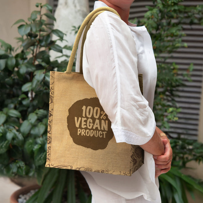 Monza Jute Tote Bag - A jute tote bag in brown, featuring a logo, held by a person wearing a white shirt, against a greenery backdrop.