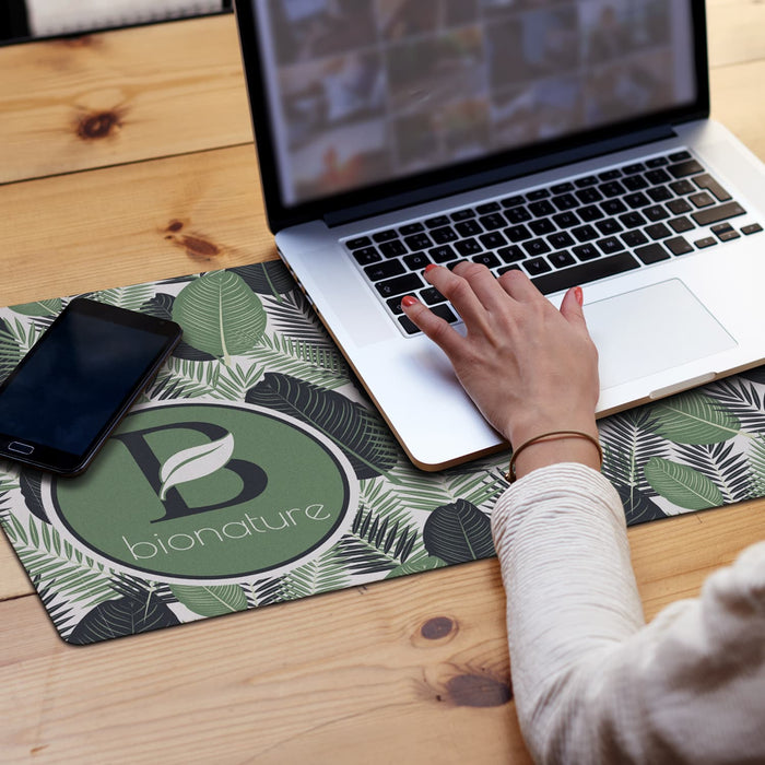 Desk Mat - A desk mat featuring a leaf pattern in green and white, with a logo design, beside a laptop and smartphone.