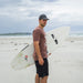 TRENDSWEAR Element Unisex T-Shirt - A man stands on a beach holding a surfboard, wearing a brown T-shirt and dark shorts.