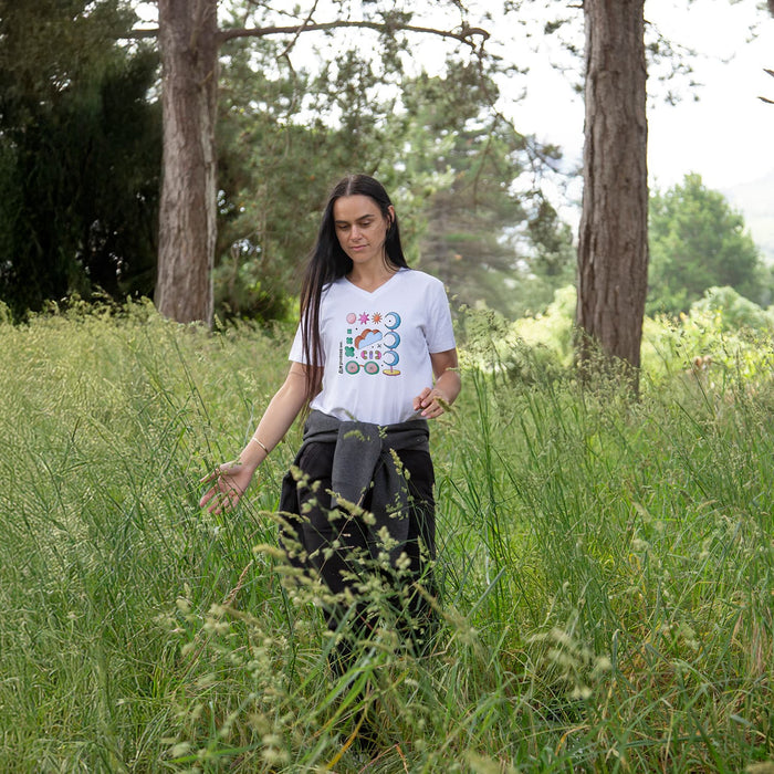 TRENDSWEAR Viva Womens T-Shirt - A woman in a white T-shirt and black pants stands in tall green grass, surrounded by trees. The T-shirt features a logo.