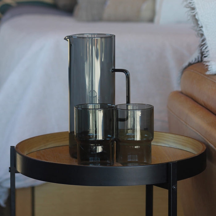 Keepsake Arlo Tumblers - Set of 4 - A drink bottle and two tumblers in smoky grey, placed on a round table beside a couch. The items feature a sleek design.