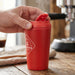 Corsica Coffee Cup - A red coffee cup with a lid, emitting steam, held by a hand on a wooden table. Features a logo.