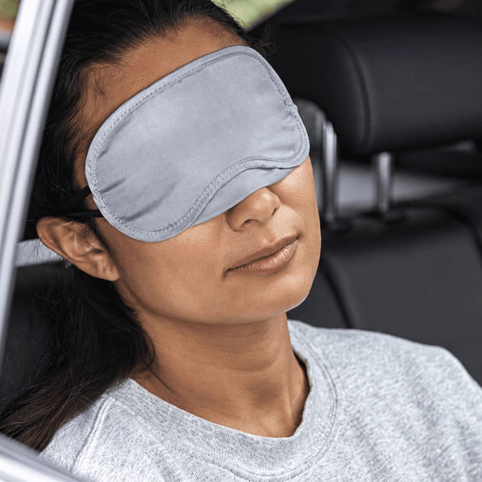 Traveler Eye Mask - A grey eye mask rests over a woman's eyes as she relaxes in a car seat, featuring a logo on the mask.