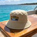 Bondi Washed Bucket Hat - A beige bucket hat with a logo sits on a wooden surface by turquoise water.