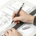 STAEDTLER The Pencil Set - A hand holding a grey pencil sketches on a blueprint with a ruler and a silver mechanical pencil nearby.