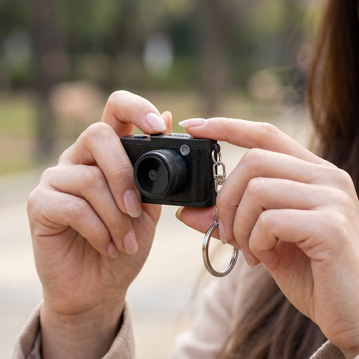 Candid Mini Camera - A compact black camera held in a person's hand, featuring a keychain attachment.