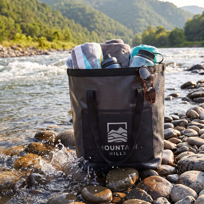Frontier Bucket Bag - A black bucket bag with towels and drink bottles sits on rocky riverbank, surrounded by a scenic landscape.