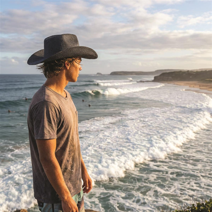 Airlie Paper Straw Hat - A person wearing a black straw hat stands by the ocean, gazing at the waves and surfers.