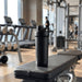 Forge Double Wall Shaker 750ml - A drink bottle with a double-wall design in black, placed on a gym bench with exercise equipment in the background.