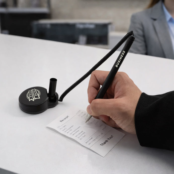 Counter Fixed Signature Pen D - A black pen with a coil attached is being used to write on a receipt at a reception desk.
