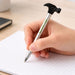 Hammer Shape Ballpoint Pen - A hammer-shaped ballpoint pen in silver and black, held over a blank notepad with lines.