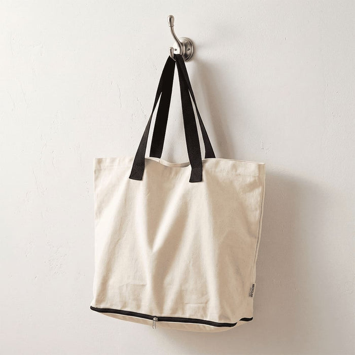 Sierra Foldable GRS Recycled  Cotton Tote Bag - A beige foldable tote bag with black straps hanging on a hook. It features a logo.