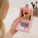 Suqare Folding Pocket Mirror - A square folding pocket mirror in pink, reflecting a person applying lipstick. Classic makeup brushes are visible in the background.