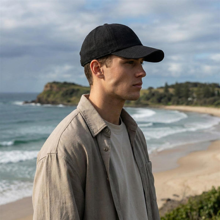 Lennox Linen Cap - A black linen cap is worn by a person on a beach, with ocean waves and cliffs in the background.