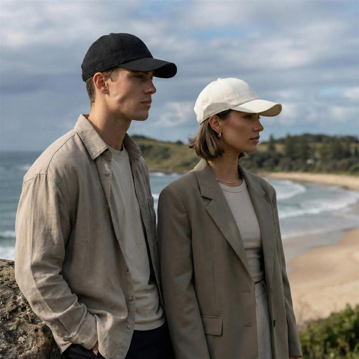 Lennox Linen Cap - A black cap and a white cap worn by a man and a woman against a coastal backdrop. The caps have logos.