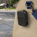 Clip Tune Magnetic Outdoor Bluetooth Speaker - A black Bluetooth speaker attached to a bag, set against an outdoor urban background. It features controls and a logo.