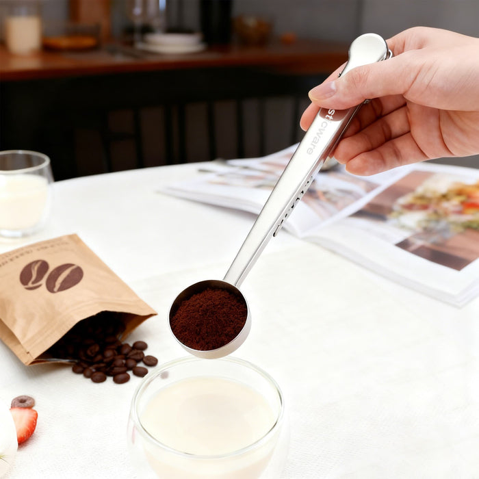 Coffee Measuring Spoon With Clip - A stainless steel coffee measuring spoon with a clip, holding ground coffee above a glass of milk on a table.