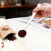 Coffee Measuring Spoon With Clip - A stainless steel coffee measuring spoon with a clip, holding ground coffee above a glass of milk on a table.