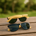 Sunglasses Bamboo (Uncoated) - Sunglasses featuring a black frame with green lenses and yellow bamboo arms, displayed on a wooden surface.