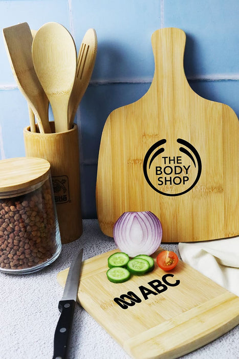 Tiga Bamboo Cutting Board - A bamboo cutting board with a handle, surrounded by kitchen utensils, sliced vegetables, and a glass container.