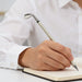 Golf Pen Gift Set - A golf club-shaped pen in silver, held by a person writing in a notebook. The setting features neutral colours.