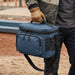 Super Duty 12 - Lunch Cooler - A blue lunch cooler bag with a handle and shoulder strap, featuring pockets and zippers, held by a gloved hand.