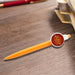 Button Badge Pen - An orange plastic pen with a circular badge on the side, resting on a wooden surface beside stationery.