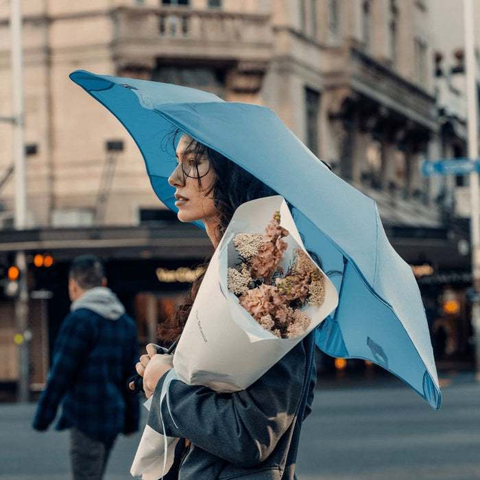 BLUNT Metro Umbrella - A blue umbrella held by a woman, who is also carrying a bouquet of flowers, in an urban setting.