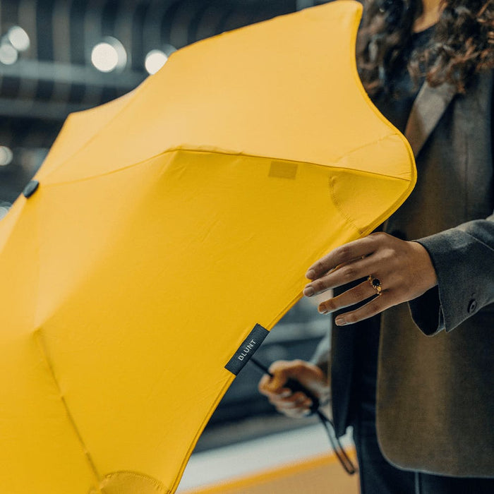 BLUNT Metro Umbrella - A yellow BLUNT Metro Umbrella is held by a person wearing a dark outfit, showcasing its sleek design.