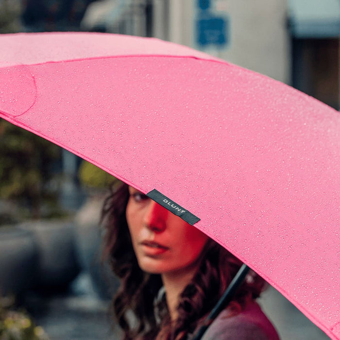 BLUNT Classic Umbrella - A pink umbrella with a black brand logo, partially covering a person, with raindrops on its surface.