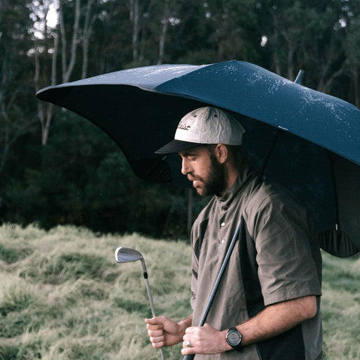 BLUNT Sport Umbrella - A black sport umbrella held by a man wearing a cap and grey shirt, standing on grass with trees in the background.