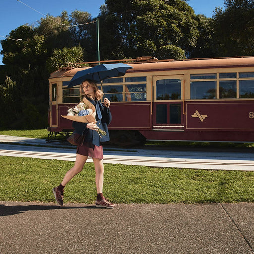BLUNT Neo Umbrella - A woman carries a BLUNT Neo Umbrella in navy blue while walking beside a vintage tram in a green outdoor setting.