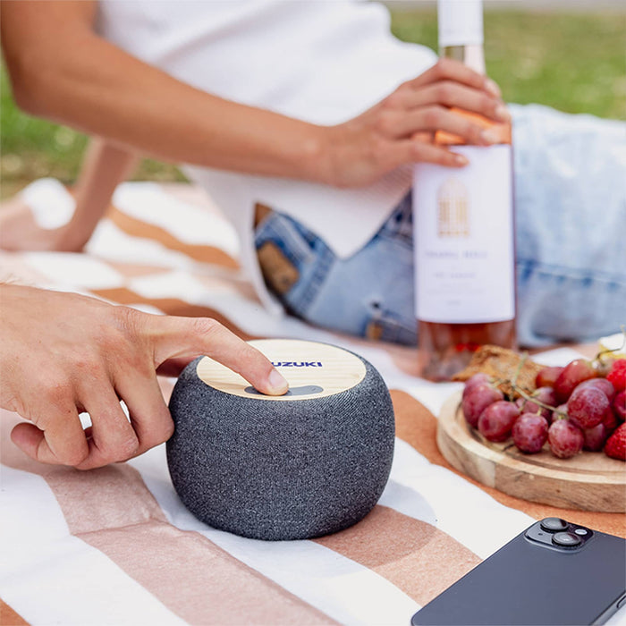 Boomer Bamboo Wireless Speaker - A circular, grey wireless speaker with a wooden top sits on a picnic blanket, alongside a bottle of wine and grapes.