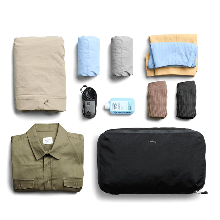 Bellroy Lite Packing Cube 12L - A black packing cube contains folded clothes, including shirts, pants, and socks, alongside a small bottle and a cable.