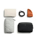 Bellroy Lite Packing Cube 6L - A cream blanket, grey quilted item, black packing cube, an orange beanie, and a black belt, all neatly arranged.