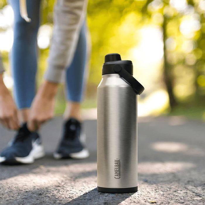 CamelBak - Thrive Chug VSS 1.2L - A drink bottle in stainless steel with a black lid, featuring a logo, set on a path surrounded by greenery.