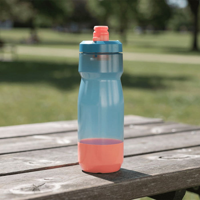 CamelBak - Podium 0.7L - A drink bottle in blue and coral, featuring a screw-top lid and a curved design, resting on a wooden table outdoors.