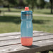 CamelBak - Podium 0.7L - A drink bottle in blue and coral, featuring a screw-top lid and a curved design, resting on a wooden table outdoors.
