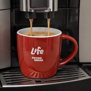 The Maxwell 330ml Ceramic Mug - A red ceramic mug with a handle, featuring a logo, is filled with coffee under a machine.