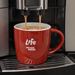 The Maxwell 330ml Ceramic Mug - A red ceramic mug with a handle, featuring a logo, is filled with coffee under a machine.