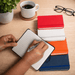 Minichroma A6 Notebook - A set of colourful notebooks in red, white, orange, and blue, with a hand writing in a black notebook. A coffee cup and glasses are nearby.