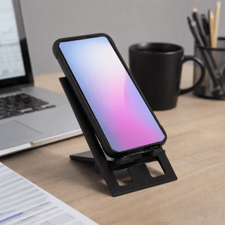 The Felix Foldable Phone Stand - A black foldable phone stand with a phone displaying a gradient screen, set on a desk with a laptop and mug.