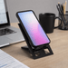 The Felix Foldable Phone Stand - A black foldable phone stand with a phone displaying a gradient screen, set on a desk with a laptop and mug.