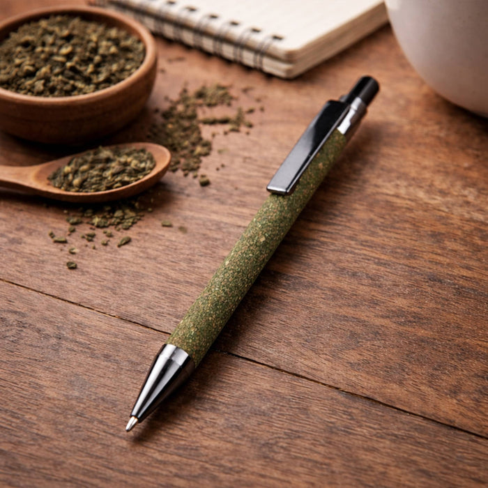 Tea Grounds Pen - A green pen with a silver tip rests on a wooden surface, surrounded by tea grounds and a notebook.