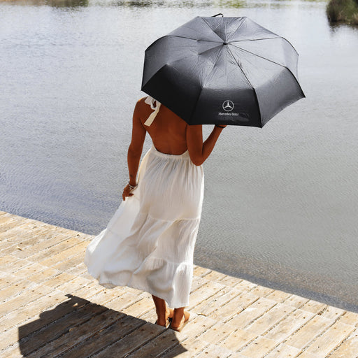 Poppins Umbrella - A black umbrella with a logo held by a person in a white dress, standing by the water.