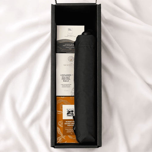 The Blunt Umbrella All Weather Set - A black umbrella is placed inside a black box alongside various snack packages. The items are neatly organised.